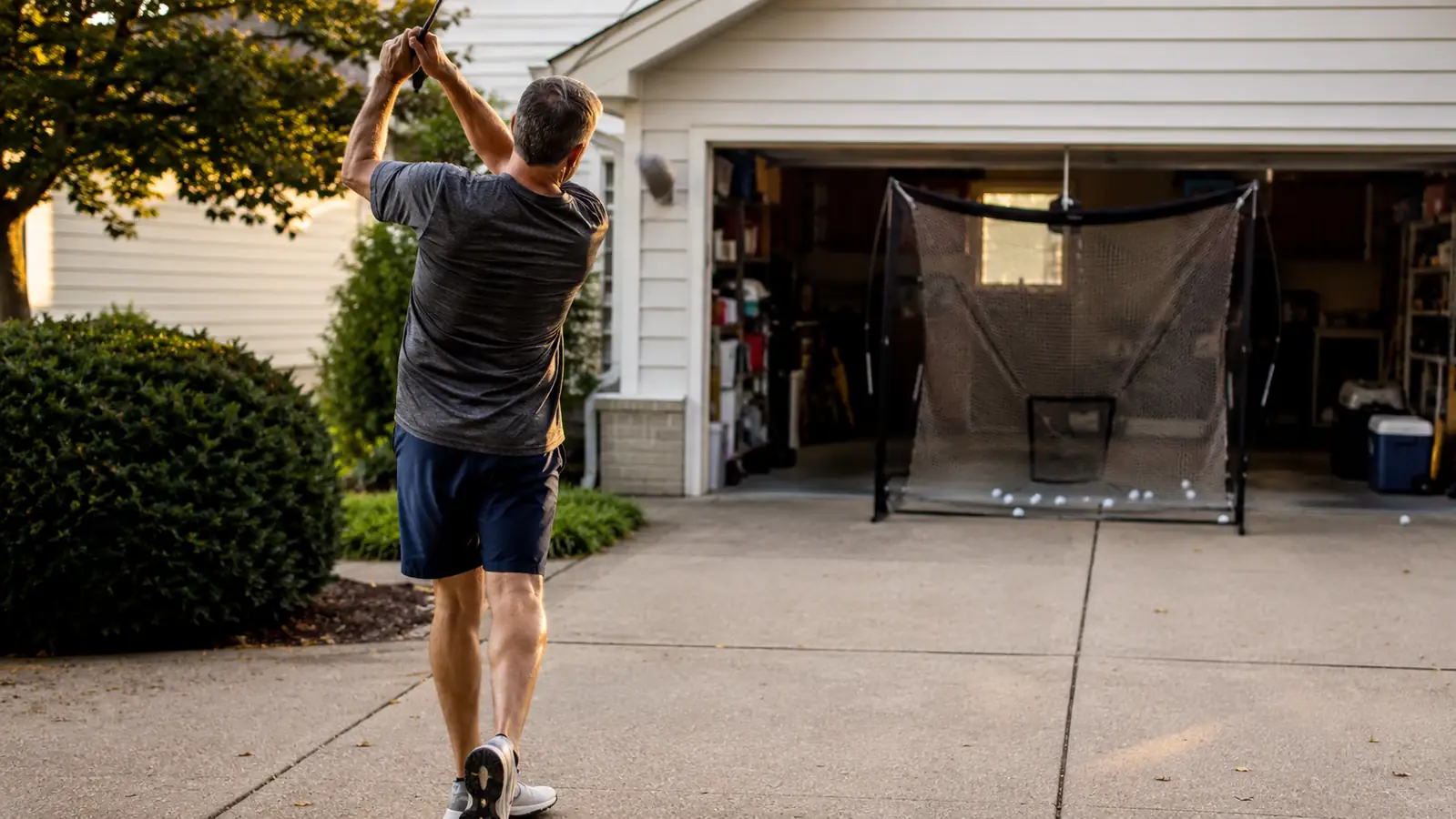 Golfer mid-swing in a driveway with a GoSports golf net set up a few feet away catching the ball