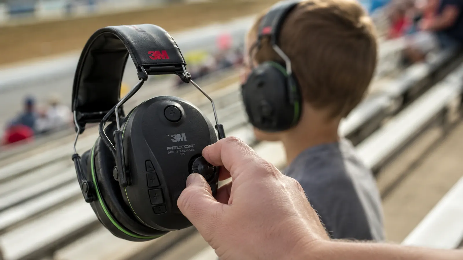 Close-up of a hand turning the volume dial on a black electronic earmuff while a boy wearing a matching pair looks on