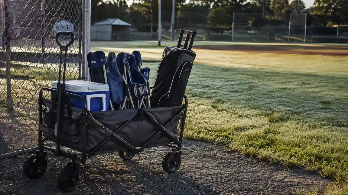 A loaded folding wagon parked at the edge of a gravel lot with a cooler, folding chairs, and a bat bag inside, ballfields in the background