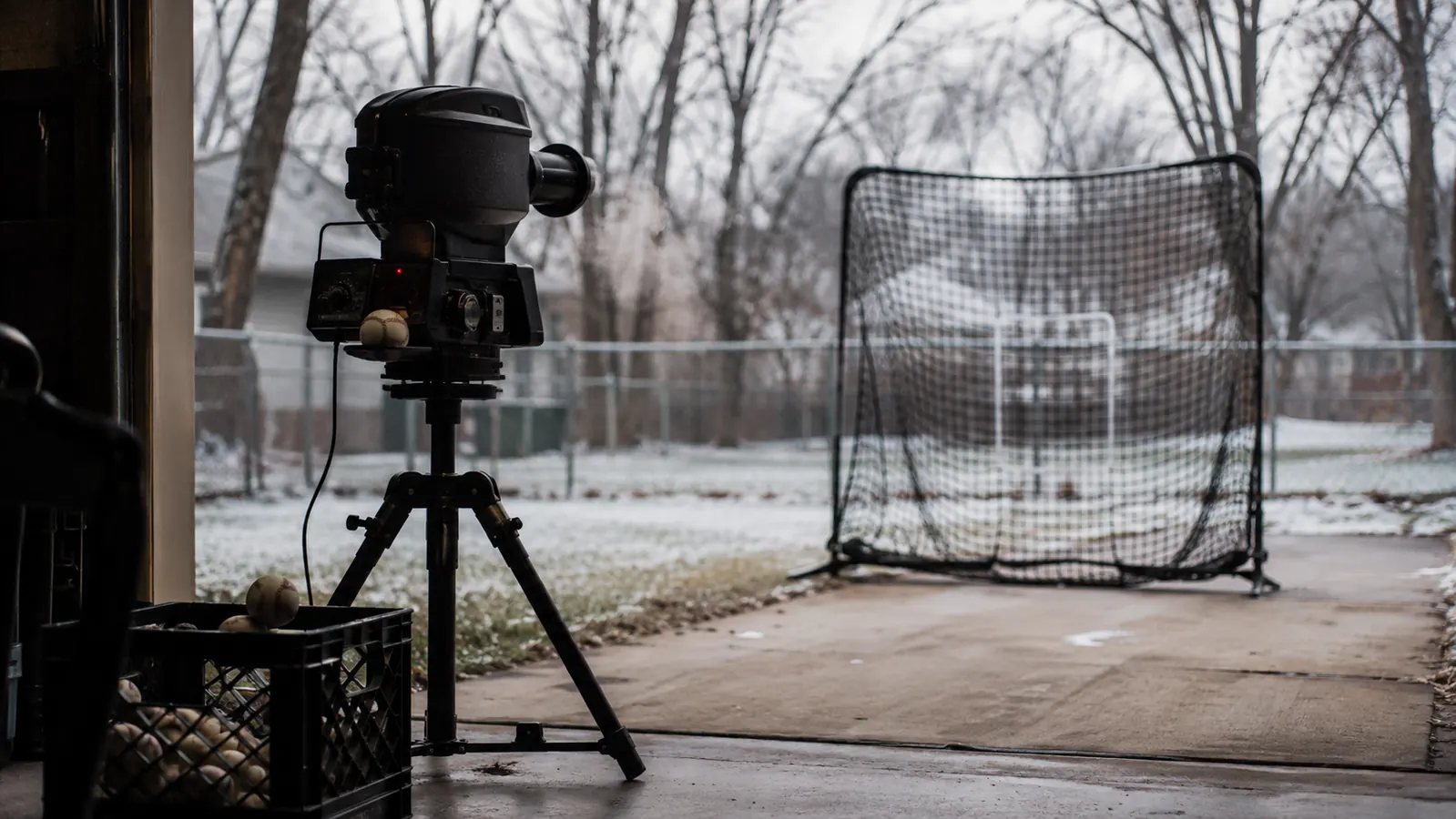 Pitching machine set up in an open garage facing a driveway batting net on a cold overcast morning