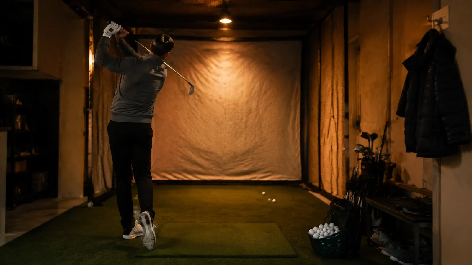 A golf practice net set up against the back wall of a garage at night, clubs leaning nearby, cold weather visible through the window
