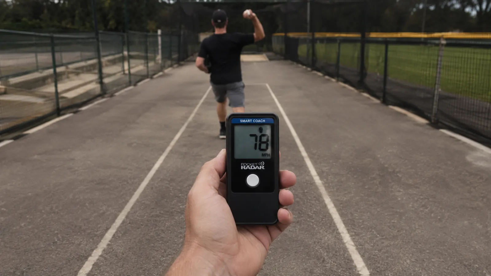 Close-up of a hand holding a Pocket Radar Smart Coach unit, aimed down the throwing lane directly behind a pitcher