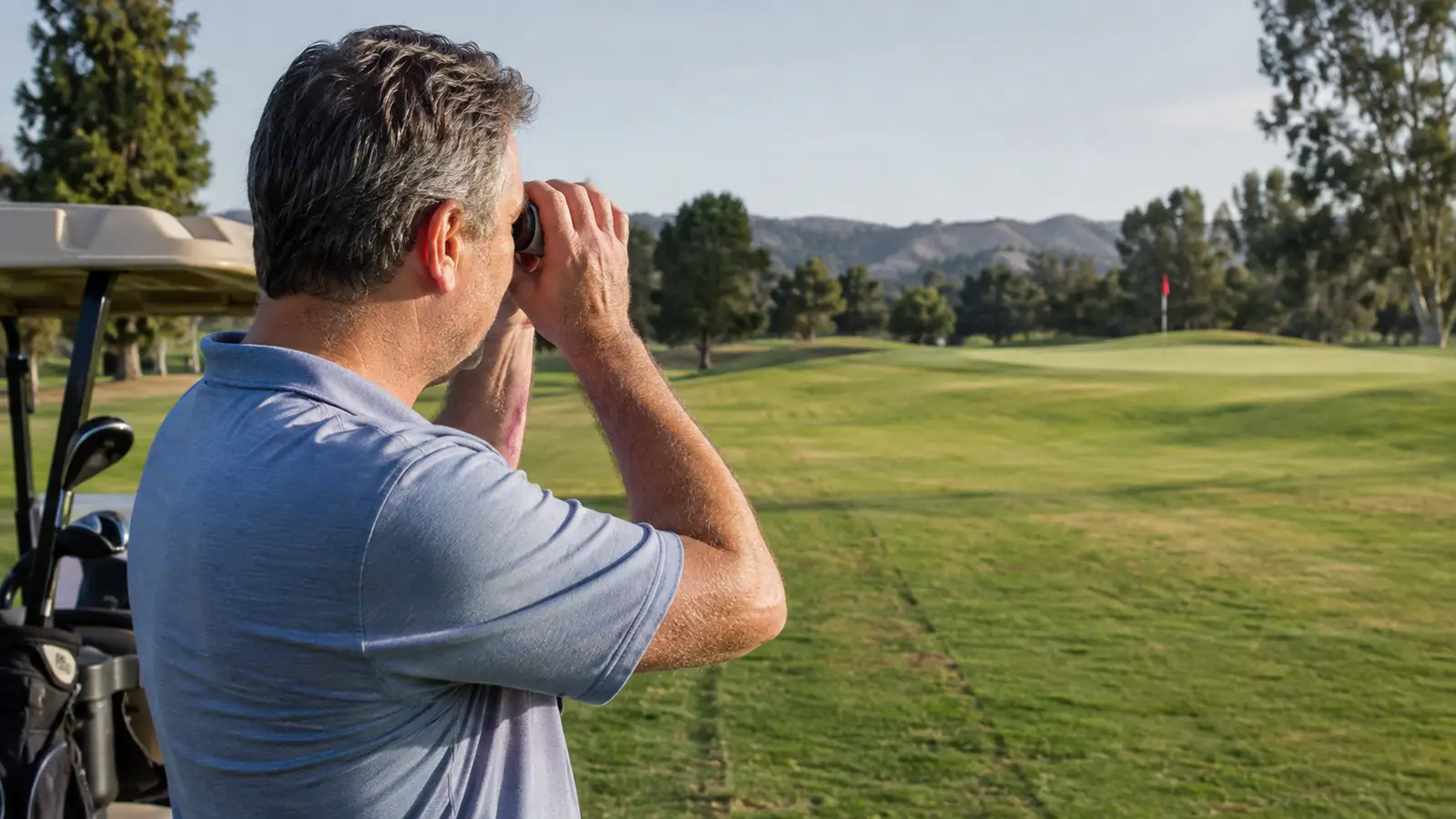 A golfer standing in the fairway holding a laser rangefinder up to his eye, aiming toward a distant flag on a sunny morning