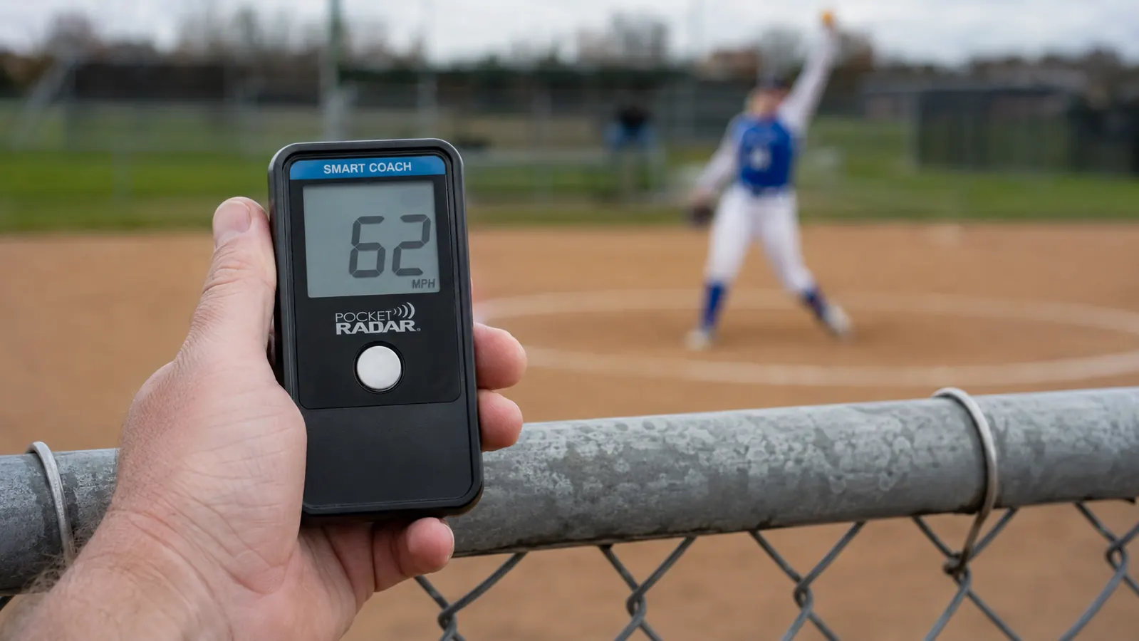 A parent's hand holding the Pocket Radar Smart Coach up behind a chain-link backstop, aimed toward a pitcher on the mound
