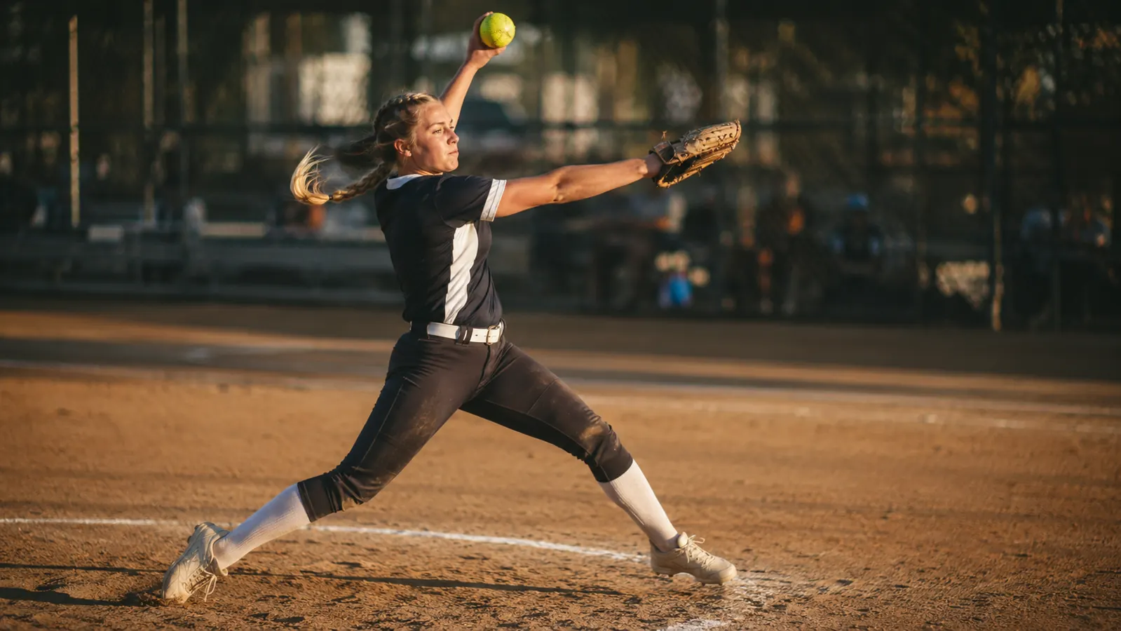 A teenage fastpitch pitcher mid windmill delivery on a sunlit softball diamond, ponytail mid-swing, focused expression