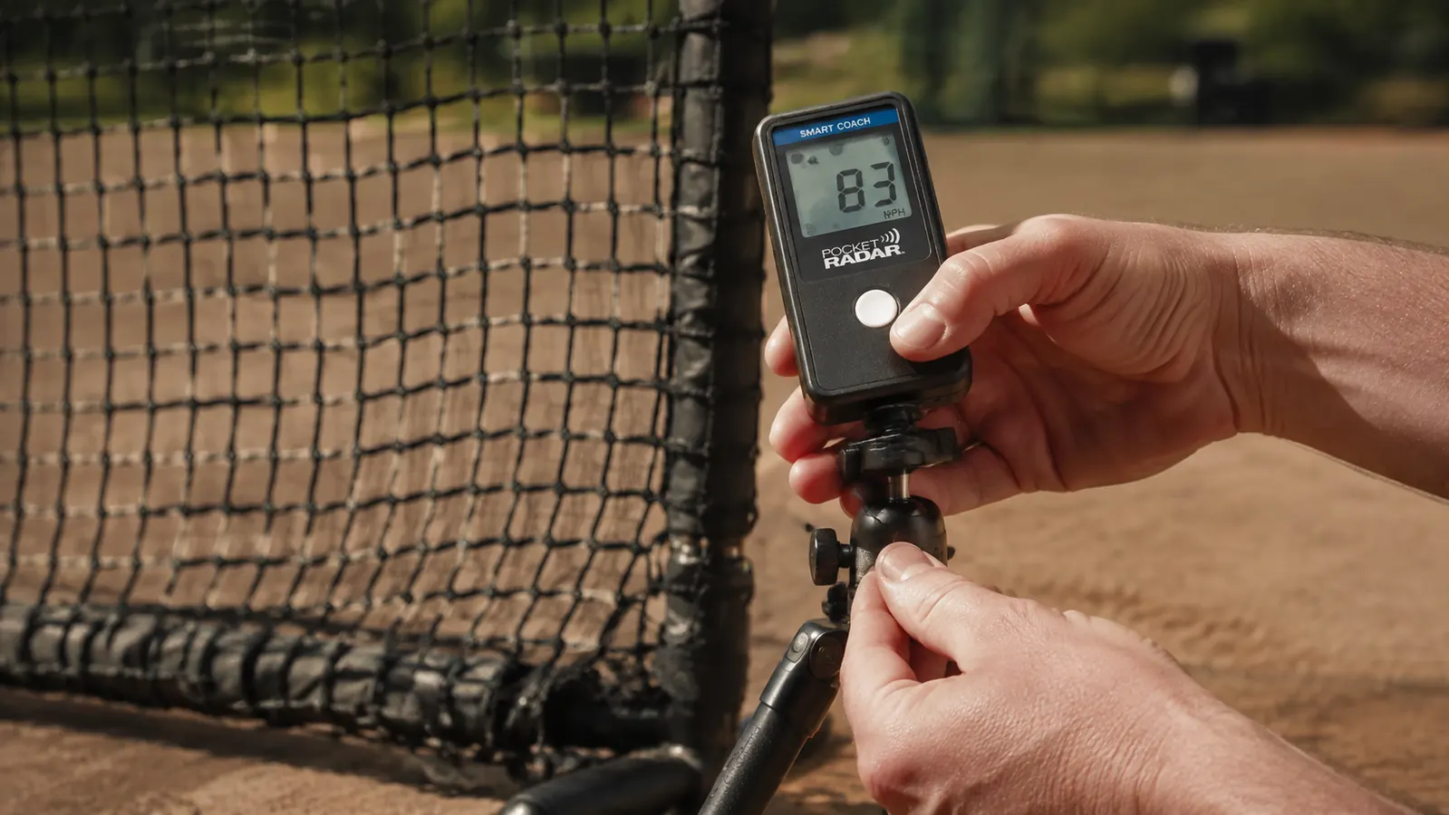 Close-up of hands mounting the Pocket Radar Smart Coach onto a small tripod behind a pitching screen