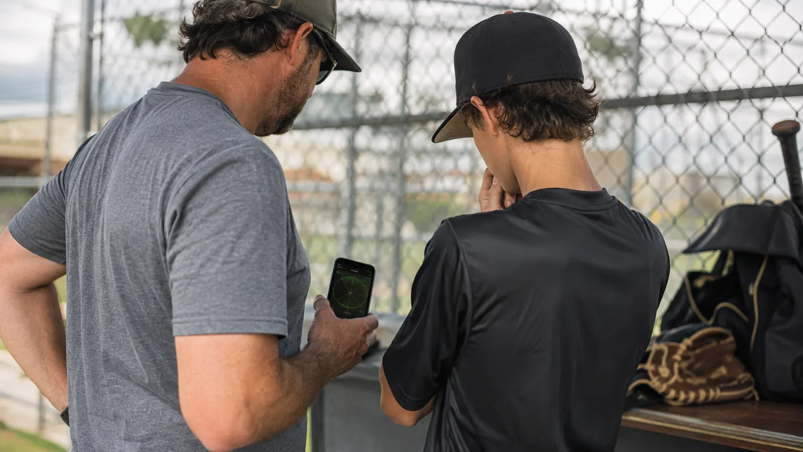 A pitcher and parent reviewing pitch velocity numbers on a phone screen dugout-side after a bullpen session