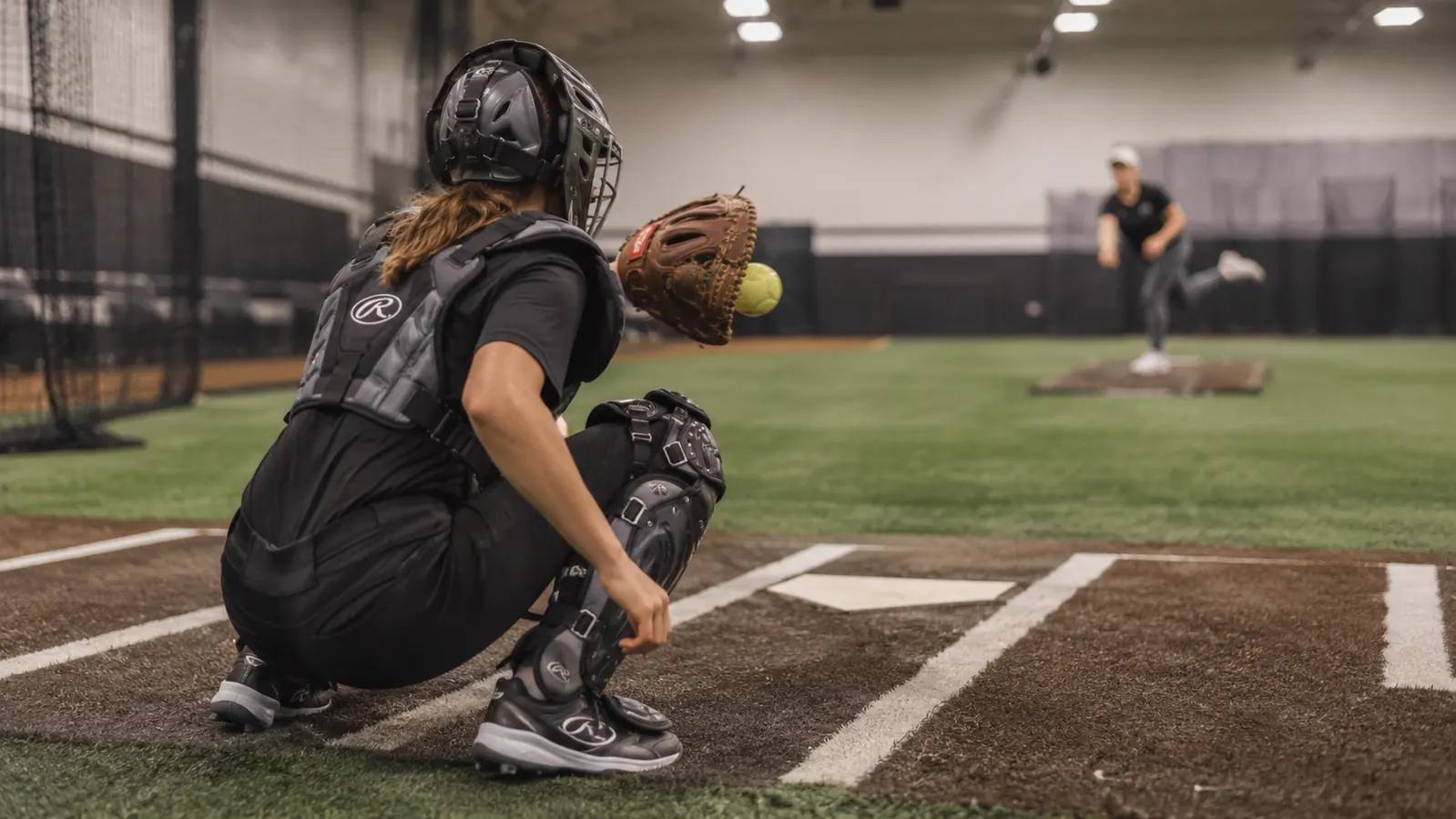 Teen fastpitch catcher crouched in full gear on indoor turf during a bullpen session