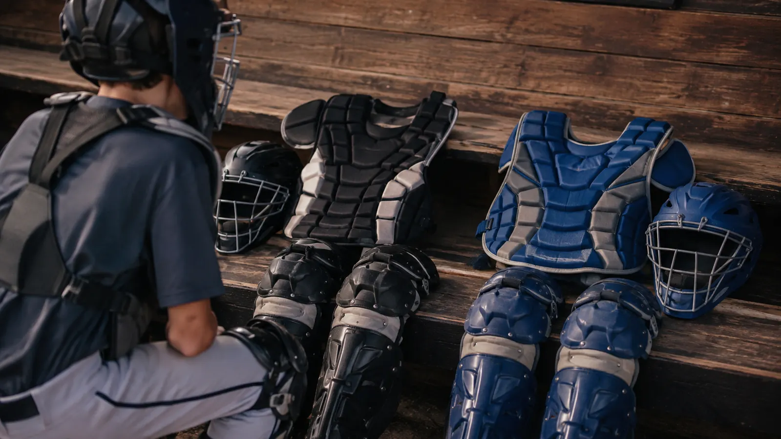 Two catcher's gear sets, one navy and red and one black and silver, laid out side by side on a dugout bench